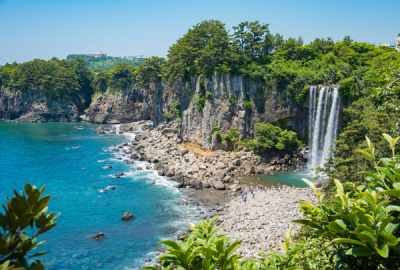 A dramatic coastal cliff face with a waterfall cascading onto a rocky shoreline on Jeju Island, South Korea, with turquoise waters and visitors exploring the beach below
