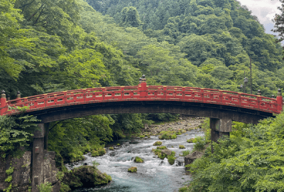 The iconic red Shinkyo Bridge arching over a rushing river surrounded by lush green forest in Nikko, Japan