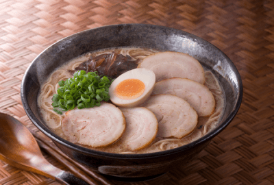 A bowl of Japanese ramen topped with chashu pork slices, a soft-boiled egg, spring onions and mushrooms, served with chopsticks and a wooden spoon