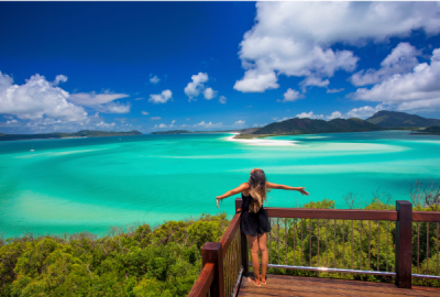 Woman standing at a lookout deck with arms outstretched over the sweeping turquoise waters and white sand of Whitehaven Beach in the Whitsunday Islands