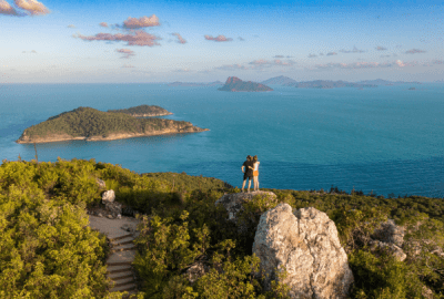 Two people embracing on a rocky summit at Passage Peak on Hamilton Island with sweeping views of turquoise Whitsunday waters and distant islands