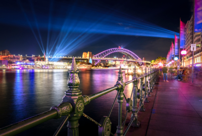 Sydney Harbour Bridge lit in violet with dramatic blue light beams radiating across the harbour waterfront during Vivid Sydney at night