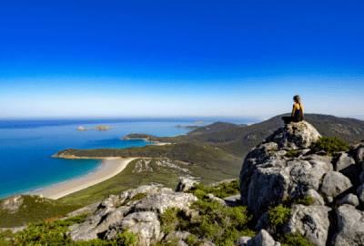 alternative text. Woman sitting on a granite rock summit at Wilsons Promontory gazing over sweeping coastal bays turquoise waters white sand beaches and green headlands
