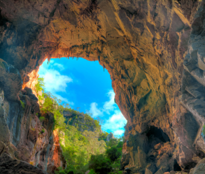 A sunlit rock cave archway at Jenolan Caves framing a lush green rainforest valley and bright blue sky in the distance