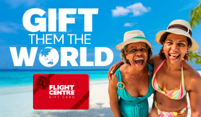 Two women laughing together on a tropical beach with turquoise water and palm trees promoting the Flight Centre Gift Card with Gift Them the World