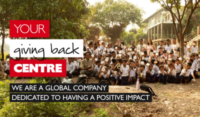 Your giving back centre - we are a global company dedicated to having a positive impact. Group of multi-racial students and teachers holding papers in the air framed by trees and a school building