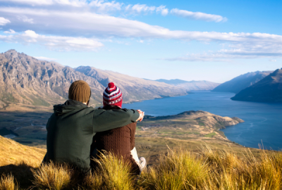 A couple sits on a tussocky hilltop overlooking a serene lake