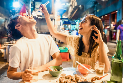 A woman holding chopsticks drops food in the mouth of a man which can best be described as culinary eroticism