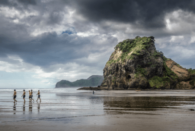 Swimmers wander across the black sands of Piha Beach at low tide with Lion Rock in the background