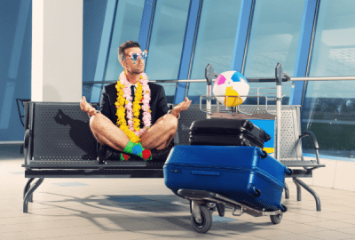 Man dressed half in business attire half in holiday clothes sits at the airport with suitcases on a trolley