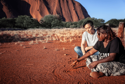 An Aboriginal woman traces stories in the sand for an onlooker beside Uluru
