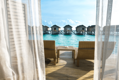 A view through thin curtains to a balcony overlooking calm, turquoise waters with arow of other overwater bungalows in the background
