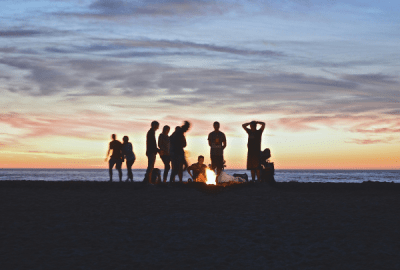 Young people gather around a beach campfire at dusk.