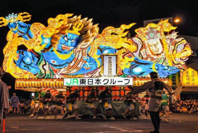 The intricate and fiery art of  an Aomori Nebuta Matsuri float lights up a dark street.