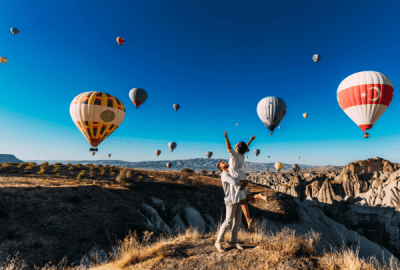 A man picks up a woman under a blue sky full of hot air balloons in Cappadocia, Turkiye