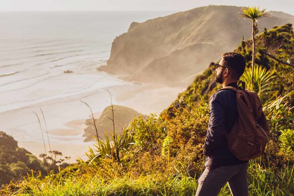 Bearded man wearing a backpack staring out at the ocean from atop a grassy cliff