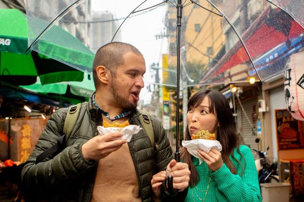 Couple eating while walking through a city street under a clear umbrella