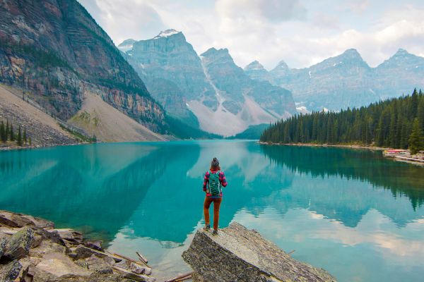 Wide shot of a person standing on a rock looking out at a large, reflective lake, pine trees and craggy mountains