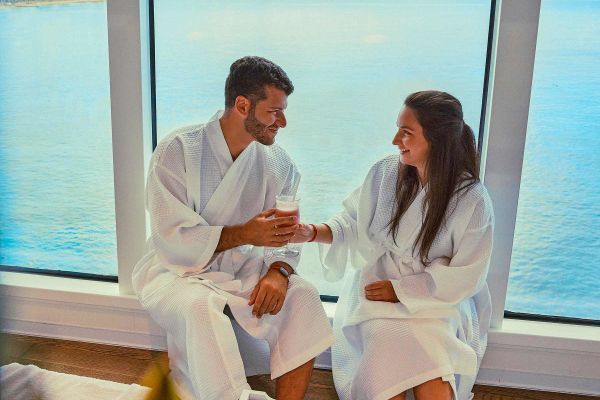 Couple in bathrobes sharing a drink sitting in front of a window on a cruise ship
