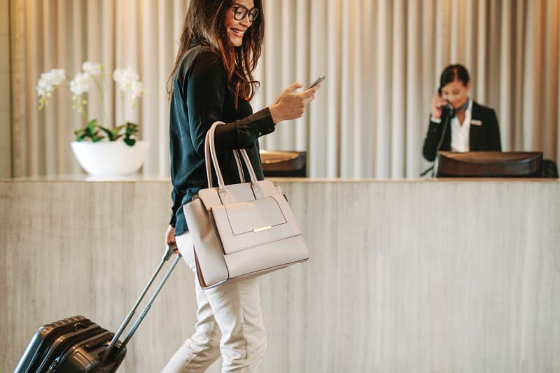 Businesswoman smiling while walking through a hotel lobby, pulling a carry-on suitcase and looking at her smartphone.