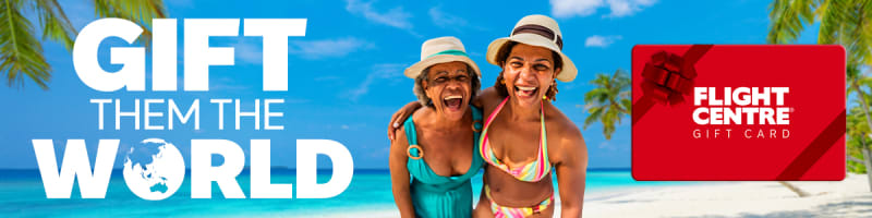Two women laughing together on a tropical beach with turquoise water and palm trees promoting the Flight Centre Gift Card with Gift Them the World