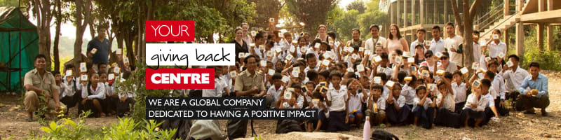 Your giving back centre - we are a global company dedicated to having a positive impact. Group of multi-racial students and teachers holding papers in the air framed by trees and a school building