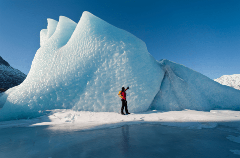 Person in an orange jacket walking beside a large ice mountain.